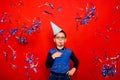 A boy in a festive cap blows into a festive pipe and stands in the rain of blue and silver confetti on a red background. Royalty Free Stock Photo