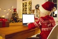 Boy in festive Santa costume and hat sitting and typing Christmas congratulation or chatting online on laptop