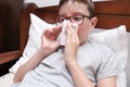 A boy with flu and fever lying in bed and blowing his nose with a paper tissue, seasonal viral diseases concept Royalty Free Stock Photo