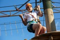 Boy in forest adventure park. Kid in orange helmet and white t shirt climbs on high rope trail. Climbing outdoor, amusement center