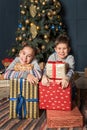 Boy and girl, brother with sister, siblings are sitting near the Christmas tree smiling happily hugging gifts boxes