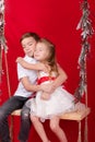 boy and girl - sister and brother - sitting on a swing decorated with christmas decor and tree branches. on red Royalty Free Stock Photo