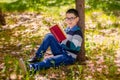 A boy with glasses is reading a book in a park while sitting on a lawn  leaning his back against a tree Royalty Free Stock Photo