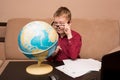 A boy in glasses and a red shirt is sitting at a black table looking at a geographical globe and doing homework, a child