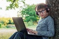 Boy in glasses sitting by tree with laptop, listen to task through headphones. Student take notes in workbook, study.