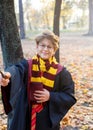 Boy in glasses stands in autumn park with gold leaves, holds book in his hands, wears in black robe. Harry Potter style