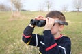 Boy, guy 7-8 years old with a feather, stalker looks through black binoculars in the park, peeps, hunts down secrets, the concept Royalty Free Stock Photo