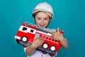 A boy in a hard hat holds a toy fire truck in his hands and smiles and shows a thumbs up. Isolated on a blue background Royalty Free Stock Photo