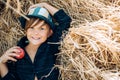 Boy in the hat holding apple and lies on the hay. Cute little child boy holding gold leaf on farm village background.