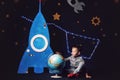 Boy in jeans and sneakers is sitting next to a toy rocket and a globe.