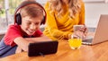 Boy In Kitchen Watching Movie On Mobile Phone Wearing Wireless Headphones As Mother Works On Laptop Royalty Free Stock Photo