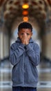 Boy with learning difficulties crying in school corridor, hands covering face, blurred background.