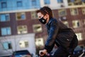 Boy in a leather jacket wearing a black hygyenic medical mask with white cross outdoors in the yard, riding a bike