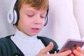 Boy is listening music in headphones in smartphone and singing a song sitting in armchair at home. Royalty Free Stock Photo