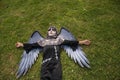 Boy in make up with white face and black eyes, dressed as an angel of death with wings, lying on his back on the grass with a lost