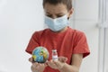 Boy in medical mask looks very seriously and holds little toy globe and ampoule with vaccine.
