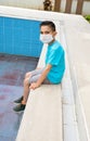 A boy with medical mask sits by an empty pool. Concept summer 2020. No tourism