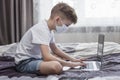 A boy in a medical mask sits in front of a laptop at home in bed. Distance learning Royalty Free Stock Photo