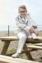 Boy with naughty face sitting on a wooden bench near the beach wears white sailor communion suit