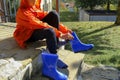 Boy in orange raincoat putting on blue rubby boots sitting on stairs in backyard. Clothing for rainy weather