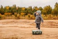 A boy in panties and a gray coat is standing on an old suitcase in the middle of a sandy country road, raising his hand to his for