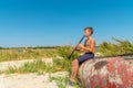 A boy is playing on a black clarinet sitting on an old wooden boat on the seashore and looking to the side, view from the side.