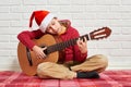 Boy playing music on guitar, dressed in a red woolen sweater and santa hat, sitting on a red checkered blanket, white brick wall o Royalty Free Stock Photo