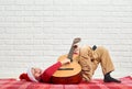 Boy playing music on guitar, dressed in a red woolen sweater and santa hat, sitting on a red checkered blanket, white brick wall o Royalty Free Stock Photo