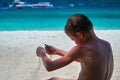 Boy playing with sand on tropical beach, run dry sand through your fingers. Rear view against the turquoise sea. Royalty Free Stock Photo
