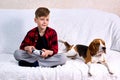 Boy plays a computer game, holds a joystick, sits in a row a puppy beagle and looks like a child playing