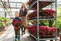Boy with a potted plant and his mom with a cart with flowers in a greenhouse.