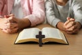 Boy praying together with his godparent at wooden table, focus on Bible and cross