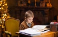 Boy reads a magic book while sitting at the table. Home interior with Christmas tree and fireplace. Traditional Royalty Free Stock Photo