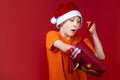 A boy on a red background in a Santa Claus hat holds a gift bag in his hands and is surprised Royalty Free Stock Photo