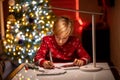 A boy in a red Christmas sweater on the background of a Christmas tree bent over the drawing in the process of decorating it with