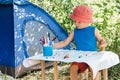 A boy in red Panama sits at a children`s table and draws markers on a white watman in nature near the camping tent