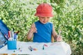 A boy in red Panama sits in nature at a children`s table and draws markers on a white watman