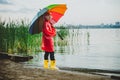 Boy in a red raincoat and yellow rubber boots stands at river bank and holding rainbow umbrella. School kid standing Royalty Free Stock Photo