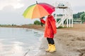 Boy in a red raincoat and yellow rubber boots stands at river bank and holding rainbow umbrella. School kid standing Royalty Free Stock Photo