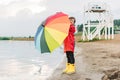 Boy in a red raincoat and yellow rubber boots stands at river bank and holding rainbow umbrella. School kid standing Royalty Free Stock Photo