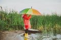 Boy in a red raincoat and yellow rubber boots stands at river bank and holding rainbow umbrella. School kid standing Royalty Free Stock Photo