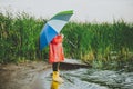 Boy in a red raincoat and yellow rubber boots stands at river bank and holding rainbow umbrella. School kid standing Royalty Free Stock Photo