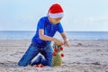 Boy in red Santa hat decorating pineapple as a Christmas tree on a sunny sandy beach by the sea