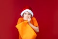 A boy in a Santa hat on a Christmas red background holds a yellow gift bag in his hands and looks fearfully into the camera Royalty Free Stock Photo