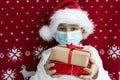 Boy in santa`s hat and face medical mask holds two gift boxes tired with ribbons on red christmas background.