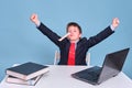 A boy in a school suit raised his hands in a victorious gesture at the computer during distance learning, copy space on a blue