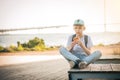 The boy sits on a bench on the waterfront with his legs crossed and eats a croissant. A teenager in a blue cap and T