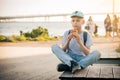 The boy sits on a bench on the waterfront with his legs crossed and eats a croissant. A teenager in a blue cap and T-shirt eats