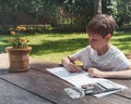 A boy sits at a wooden table in the backyard of the house and draws flowers in a pot Royalty Free Stock Photo