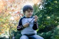 Boy sitting on green grass playing with wooden stick with blurry colourful of Autumn leaves, Smile Kid holding twig enjoy relaxing Royalty Free Stock Photo